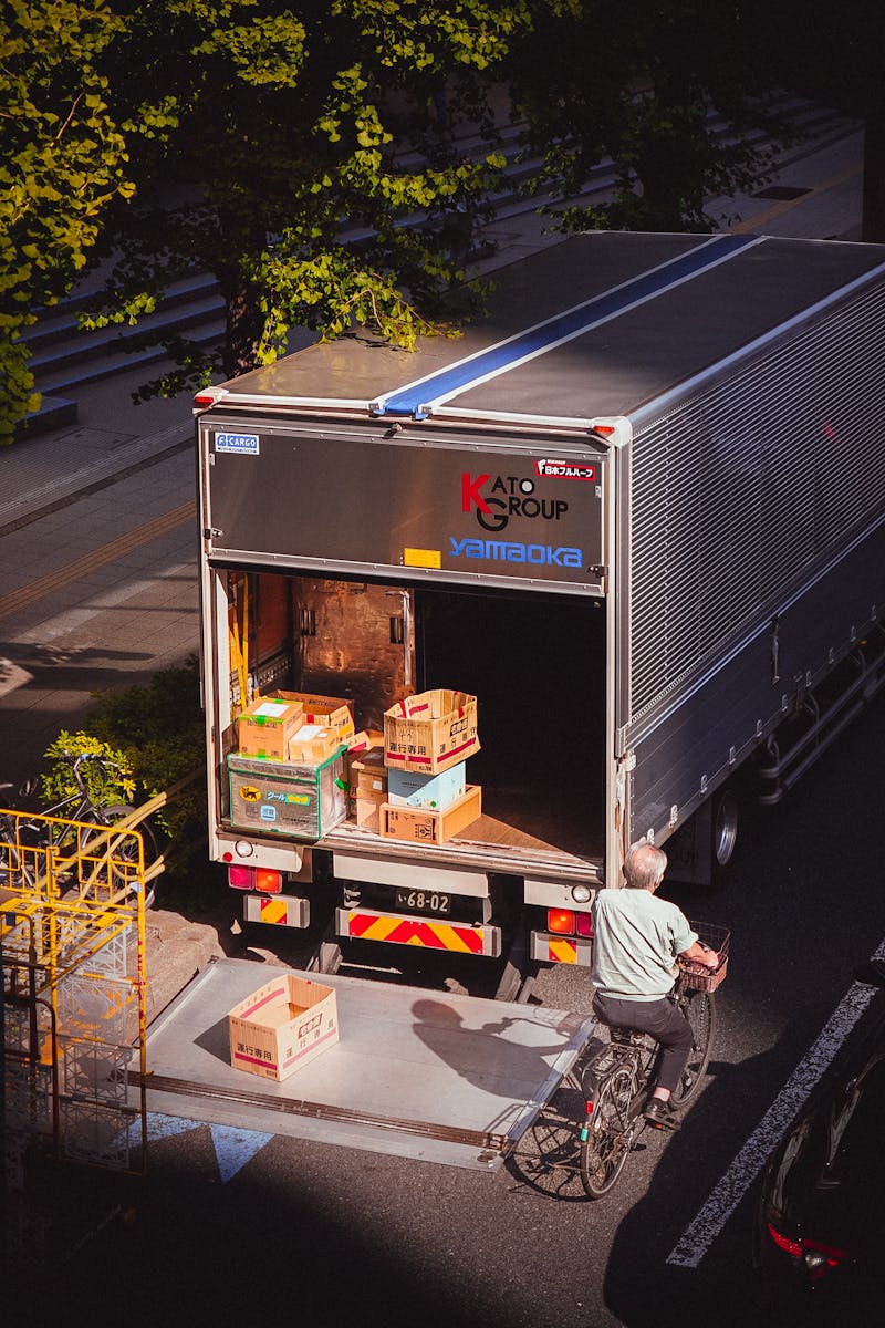 A delivery truck unloading boxes in a city street, with a bicyclist passing by on a sunny day.
